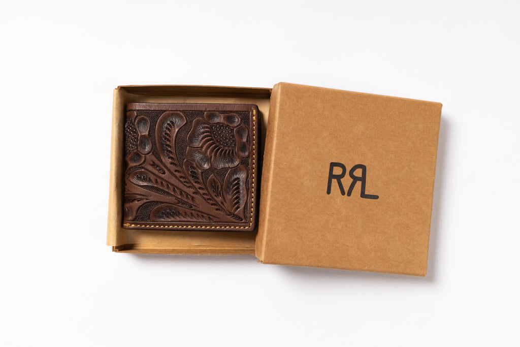 An embossed brown leather wallet with floral tooling, presented in an open kraft paper gift box with its lid showing the black monogram RRL