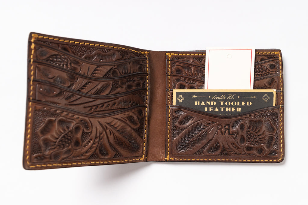 An open brown tooled leather bifold wallet with floral and scroll embossed patterns, tan contrast stitching, multiple card slots, and a branded card holder pocket reading 'Hand Tooled Leather.'.
