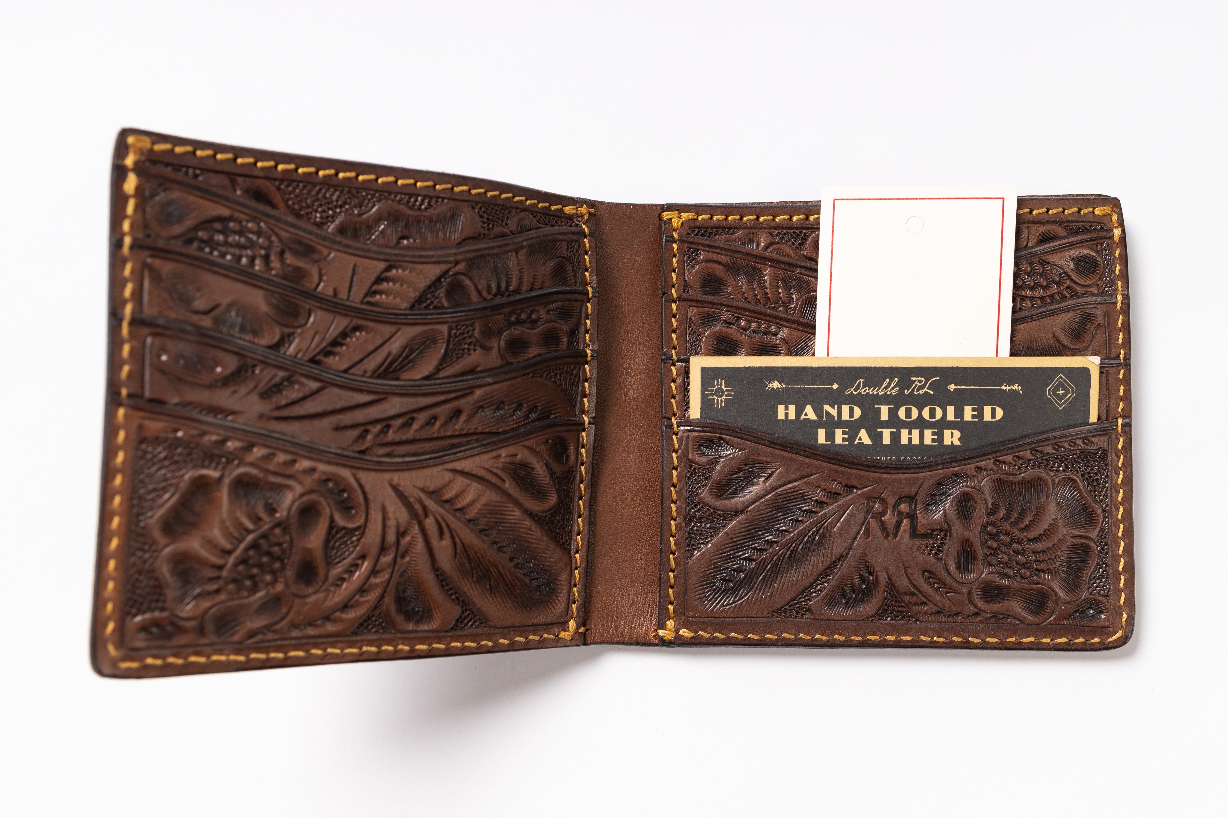 An open brown tooled leather bifold wallet with floral and scroll embossed patterns, tan contrast stitching, multiple card slots, and a branded card holder pocket reading 'Hand Tooled Leather.'.