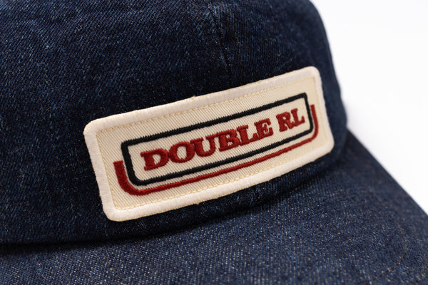 Close-up of a dark blue denim baseball cap featuring a rectangular embroidered patch on the front with the text 'DOUBLE RL' in red and black on a cream background.