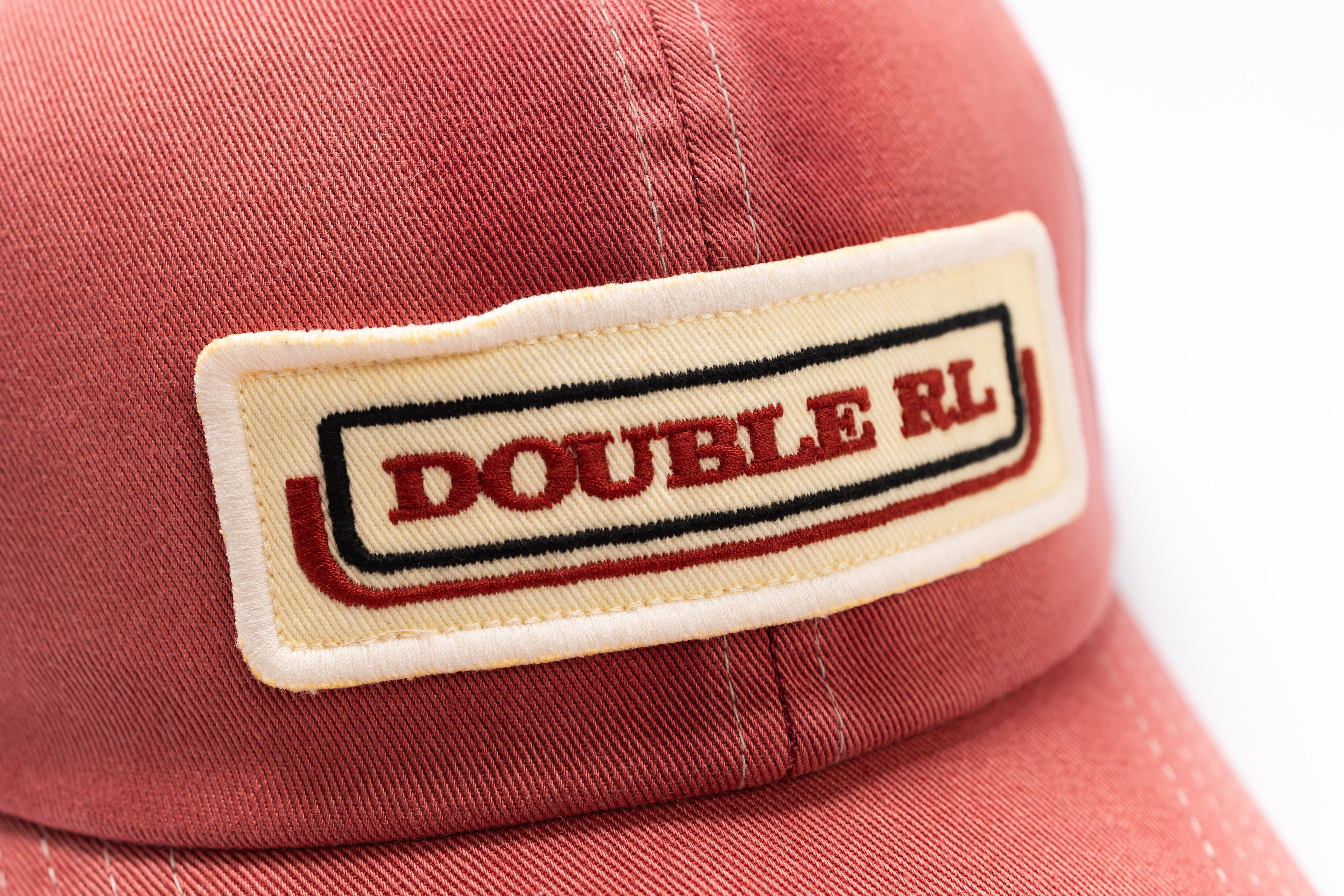 A close-up shot of a coral red baseball cap featuring a rectangular embroidered patch on the front with the text 'DOUBLE RL' in red, outlined with black and white borders.
