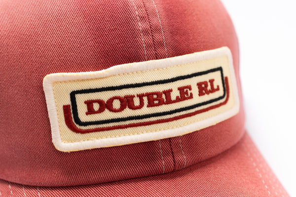 A close-up shot of a coral red baseball cap featuring a rectangular embroidered patch on the front with the text 'DOUBLE RL' in red, outlined with black and white borders.