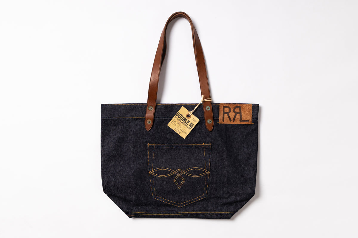 A dark indigo denim tote bag with brown leather shoulder straps, featuring a front pocket with contrast stitching and a leather patch with the letters 'RRL'.
