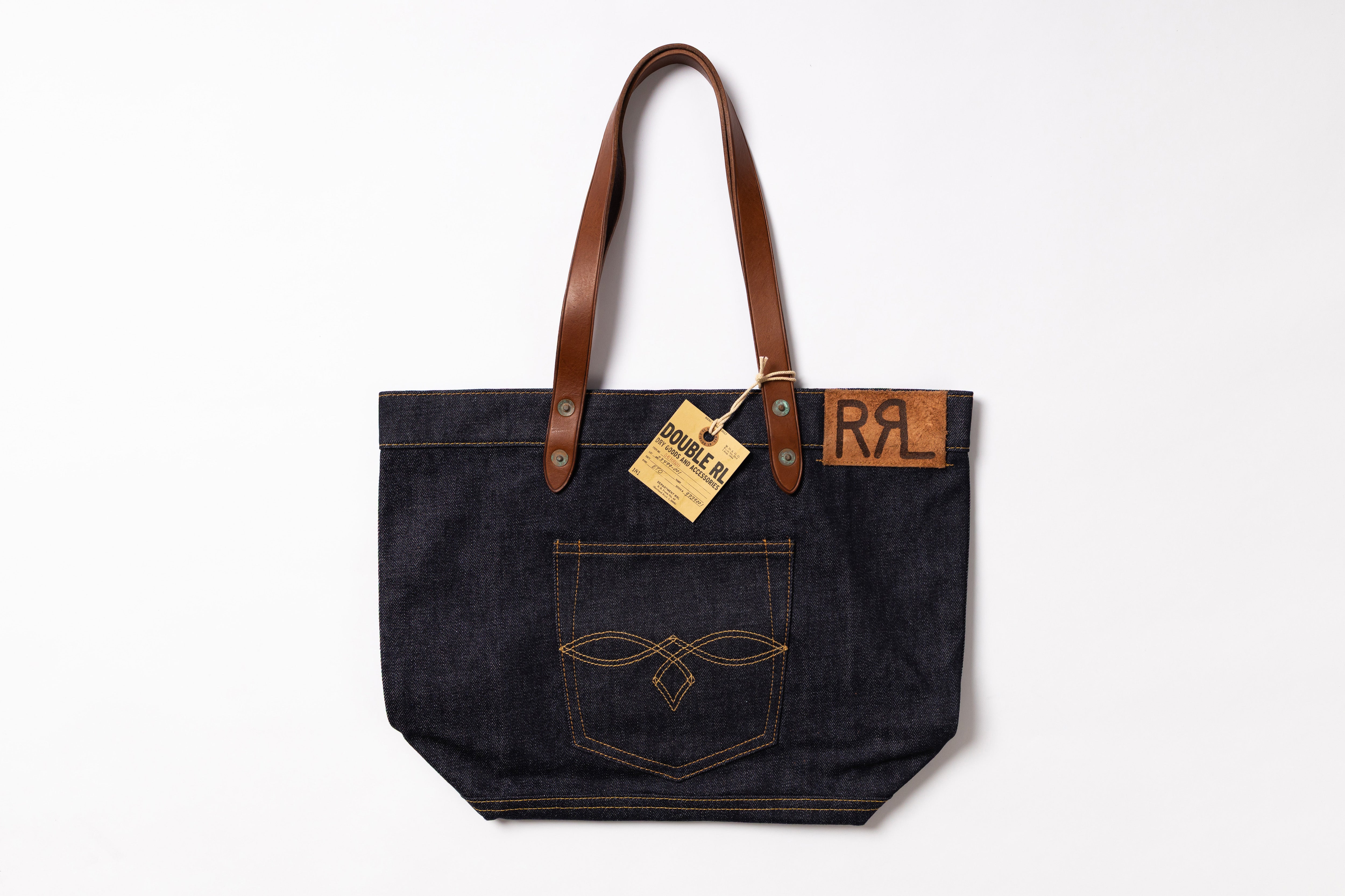 A dark indigo denim tote bag with brown leather shoulder straps, featuring a front pocket with contrast stitching and a leather patch with the letters 'RRL'.