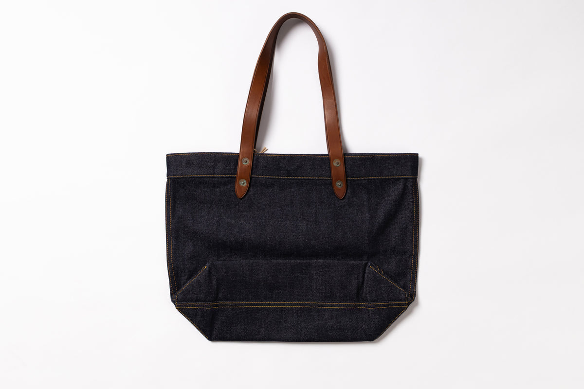 A dark indigo denim tote bag with brown leather shoulder straps, shown against a plain white background.