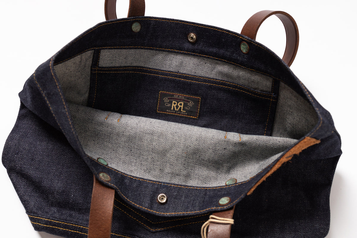 Open dark indigo denim bag showing the interior lining, brown leather handles, and a central brand patch reading RRL.