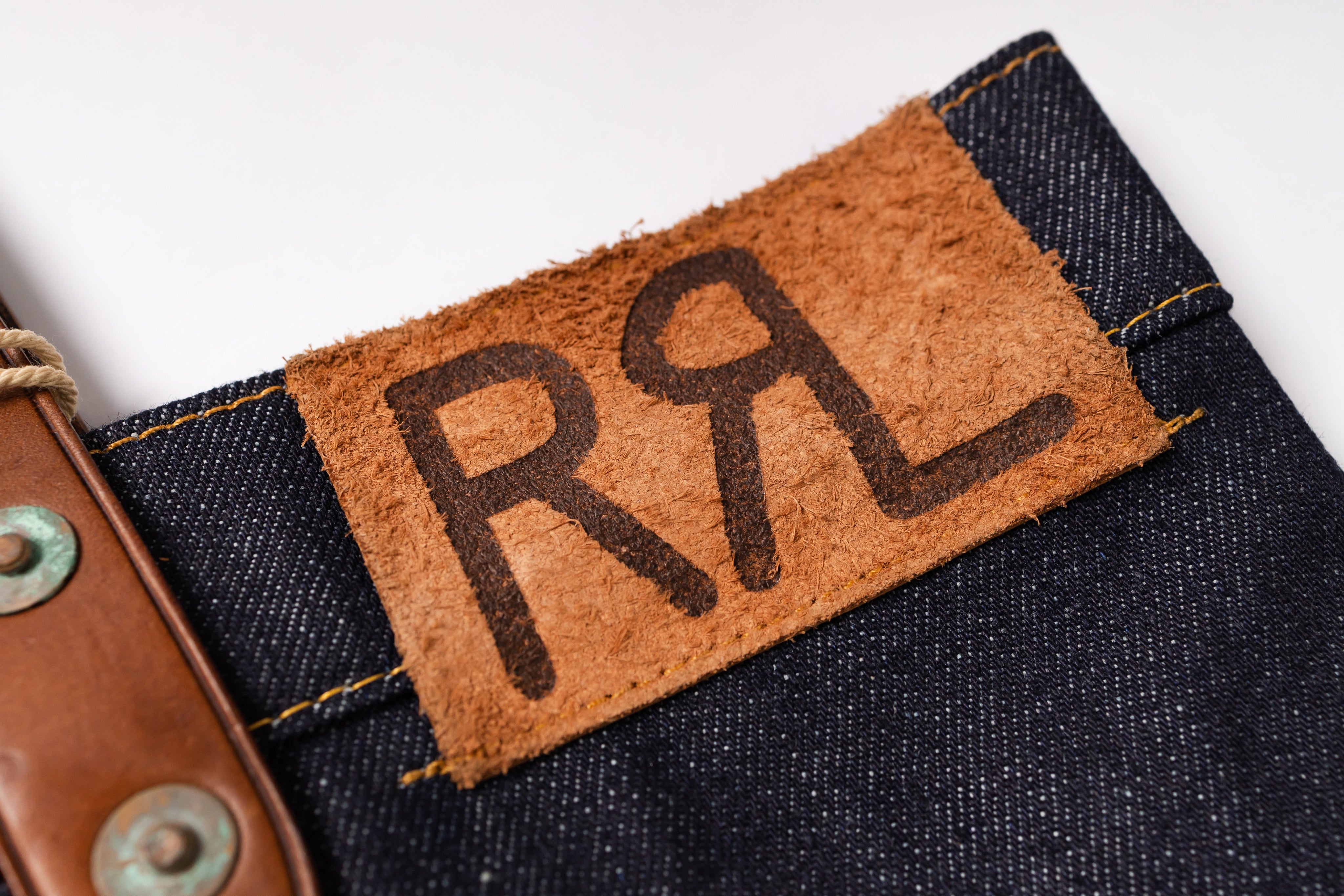 Close-up of a dark indigo denim waistband with a tan leather patch featuring the stylized initials 'RRL'.