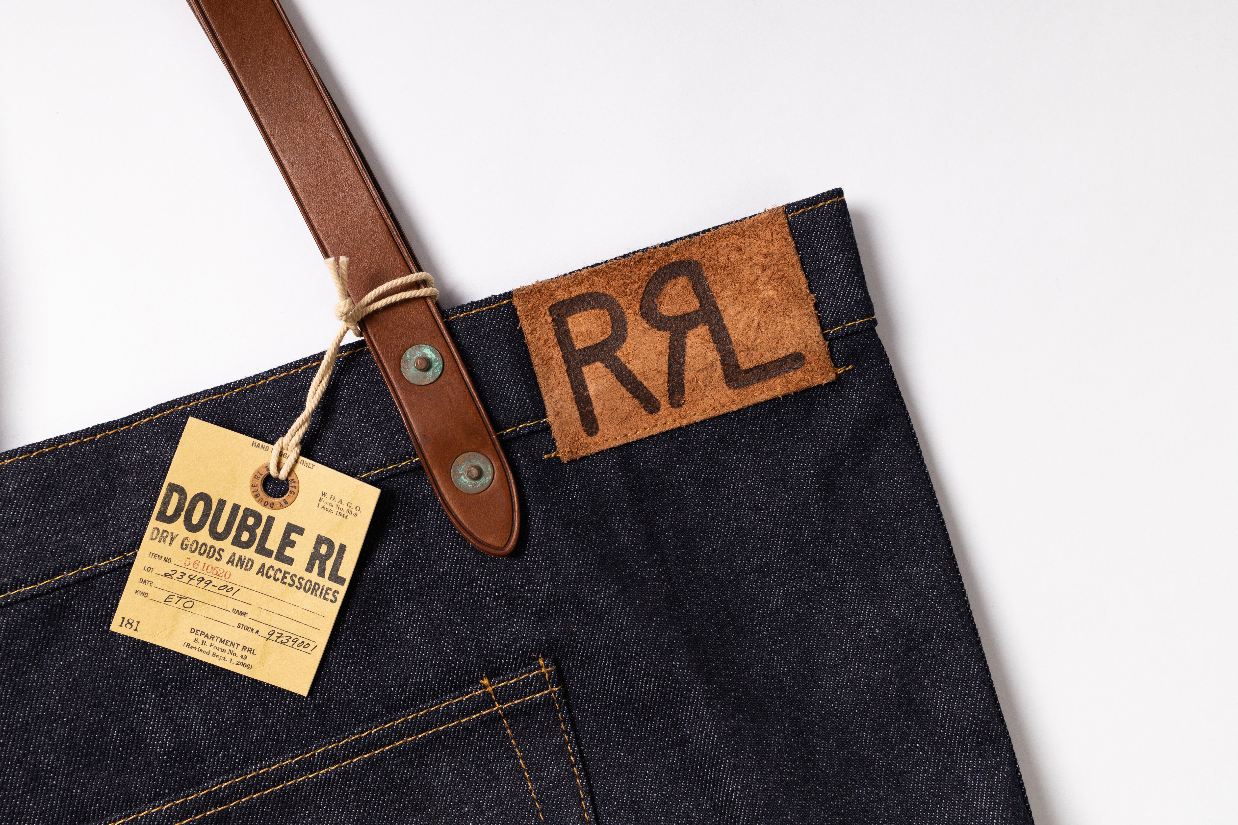 Close-up of a dark indigo denim garment, likely an apron or bib, with a brown leather strap and a tan paper brand tag reading DOUBLE RL. A matching leather patch on the garment shows the stylized 'RL' logo.