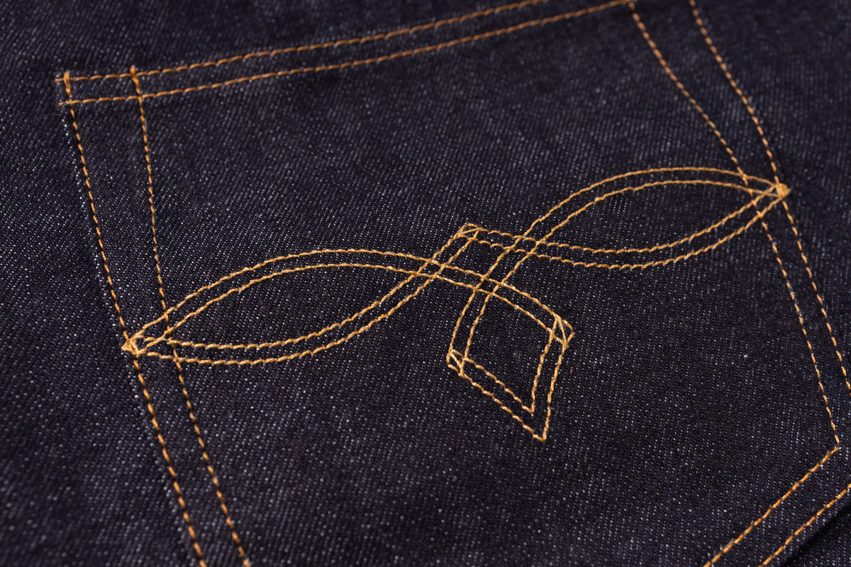 Close-up of dark indigo denim fabric with contrasting tan stitching forming a decorative leaf-like emblem on the back pocket.