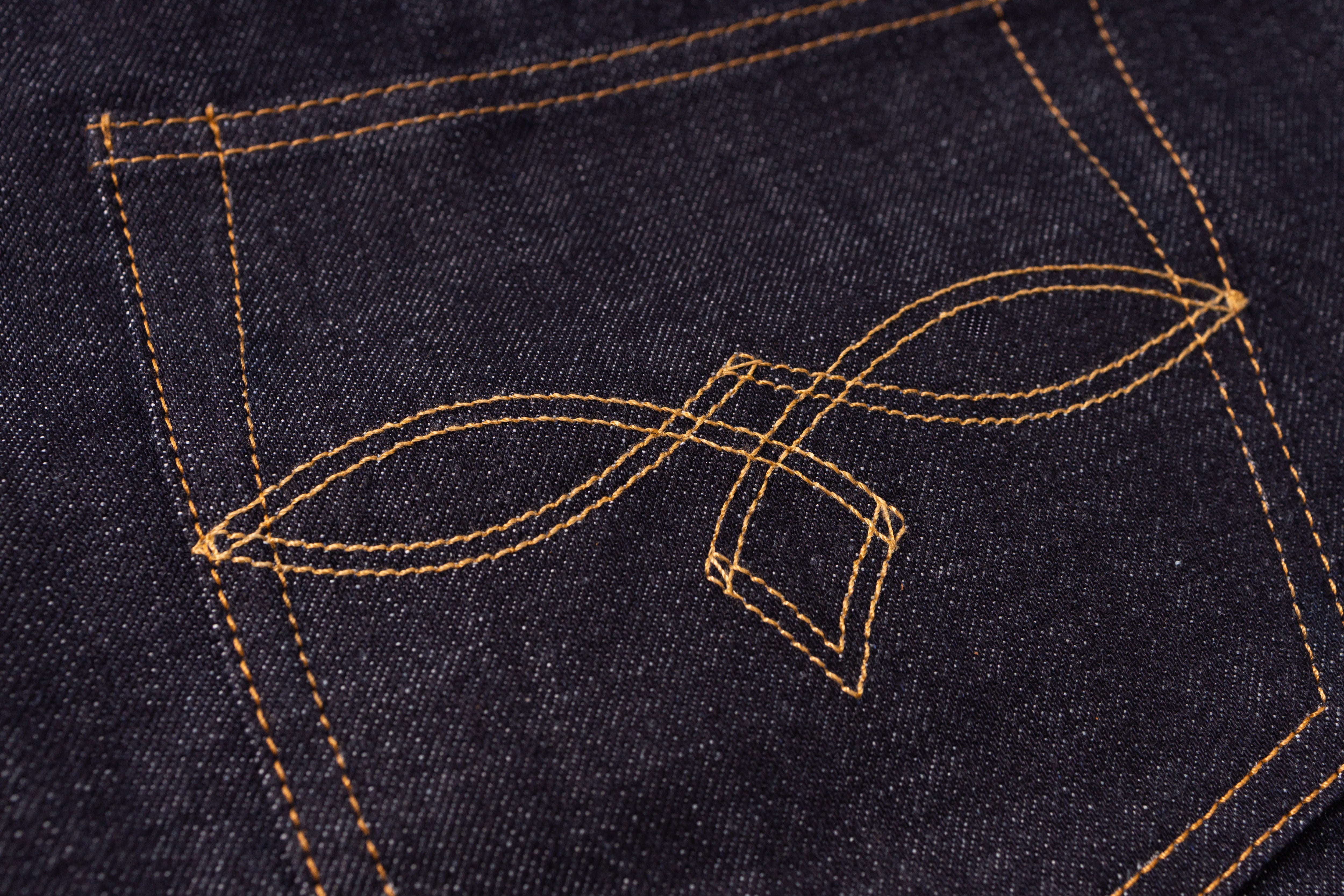 Close-up of dark indigo denim fabric with contrasting tan stitching forming a decorative leaf-like emblem on the back pocket.