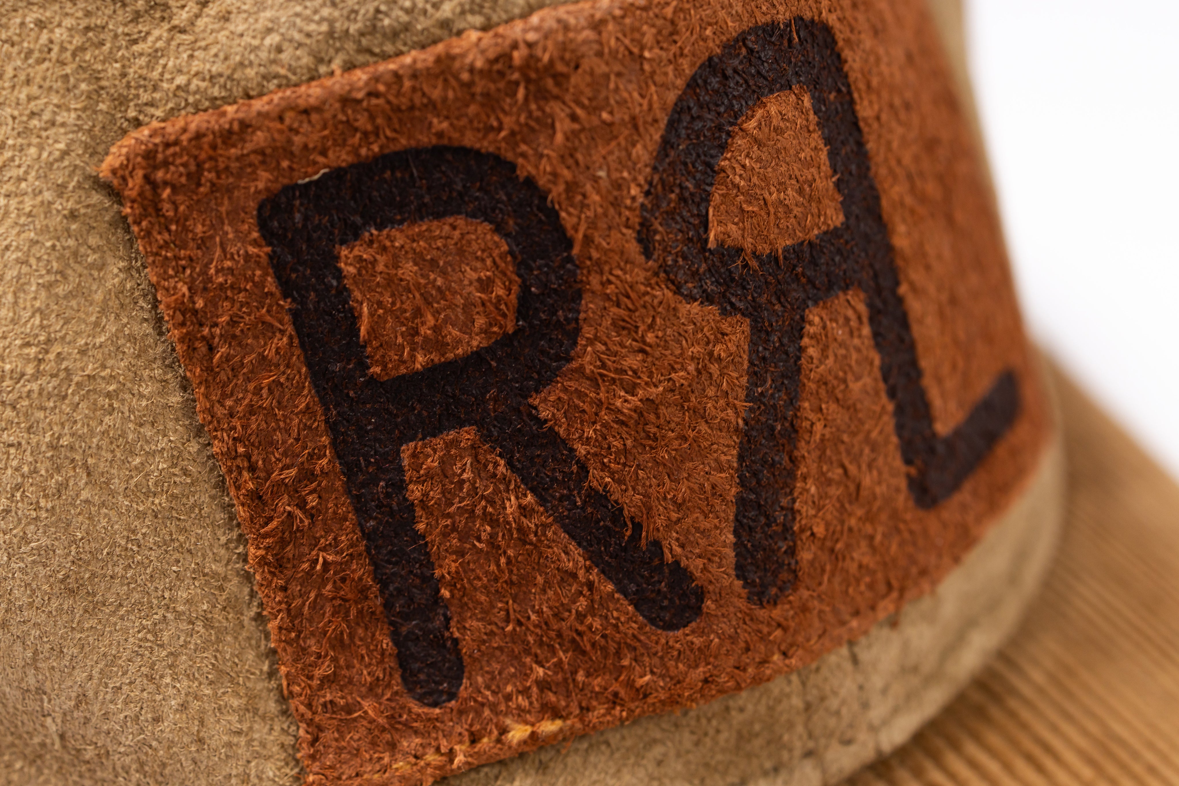 Close-up of a tan suede or leather hat with a brown patch featuring large black embroidered letters RRL
