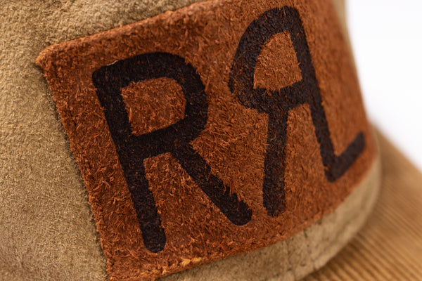 Close-up of a tan suede or leather hat with a brown patch featuring large black embroidered letters RRL