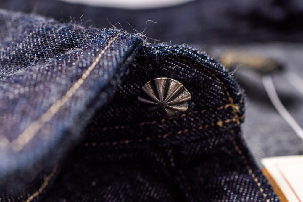 Close-up of dark indigo denim fabric with contrast tan stitching and a metallic fan-shaped rivet near the collar.