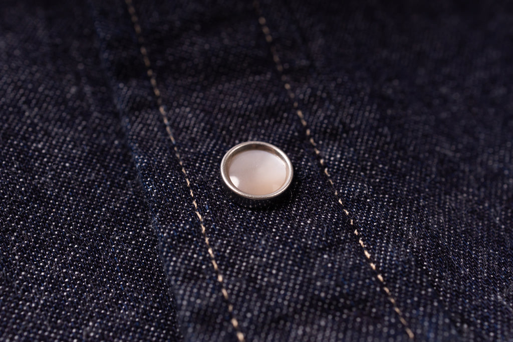 A close-up of a round silver-toned metal snap button with a pale pinkish-beige center, set onto dark indigo denim fabric with visible stitching.
