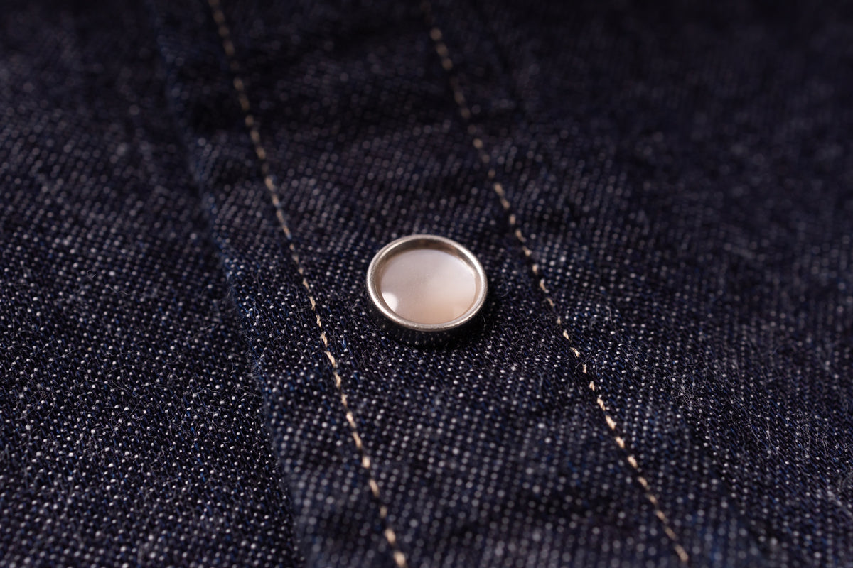 A close-up of a round silver-toned metal snap button with a pale pinkish-beige center, set onto dark indigo denim fabric with visible stitching.