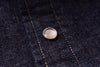 A close-up of a round silver-toned metal snap button with a pale pinkish-beige center, set onto dark indigo denim fabric with visible stitching.