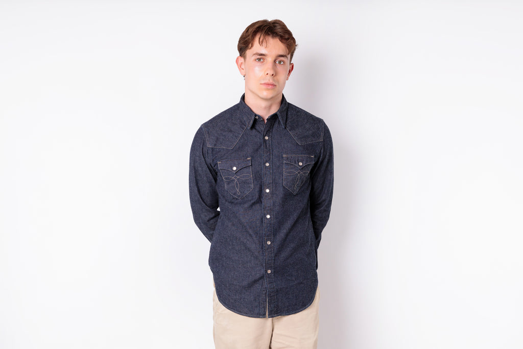 A young man wearing a dark indigo denim long-sleeve button-up shirt with two chest pockets and beige pants, standing against a plain white studio background.