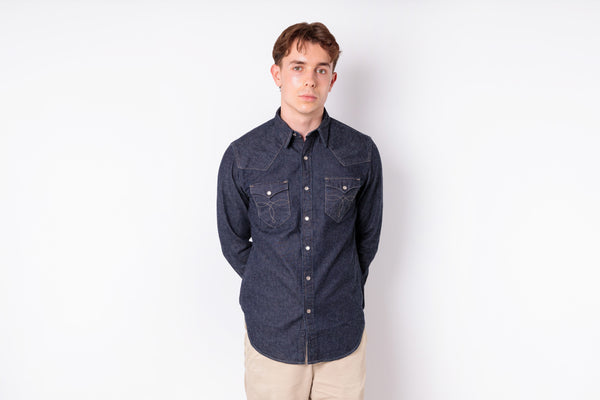 A young man wearing a dark indigo denim long-sleeve button-up shirt with two chest pockets and beige pants, standing against a plain white studio background.