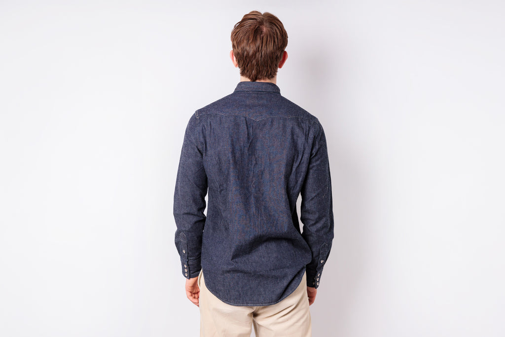 A man viewed from behind wearing a dark navy blue patterned long-sleeve button-up shirt and beige trousers, standing against a plain white studio background.