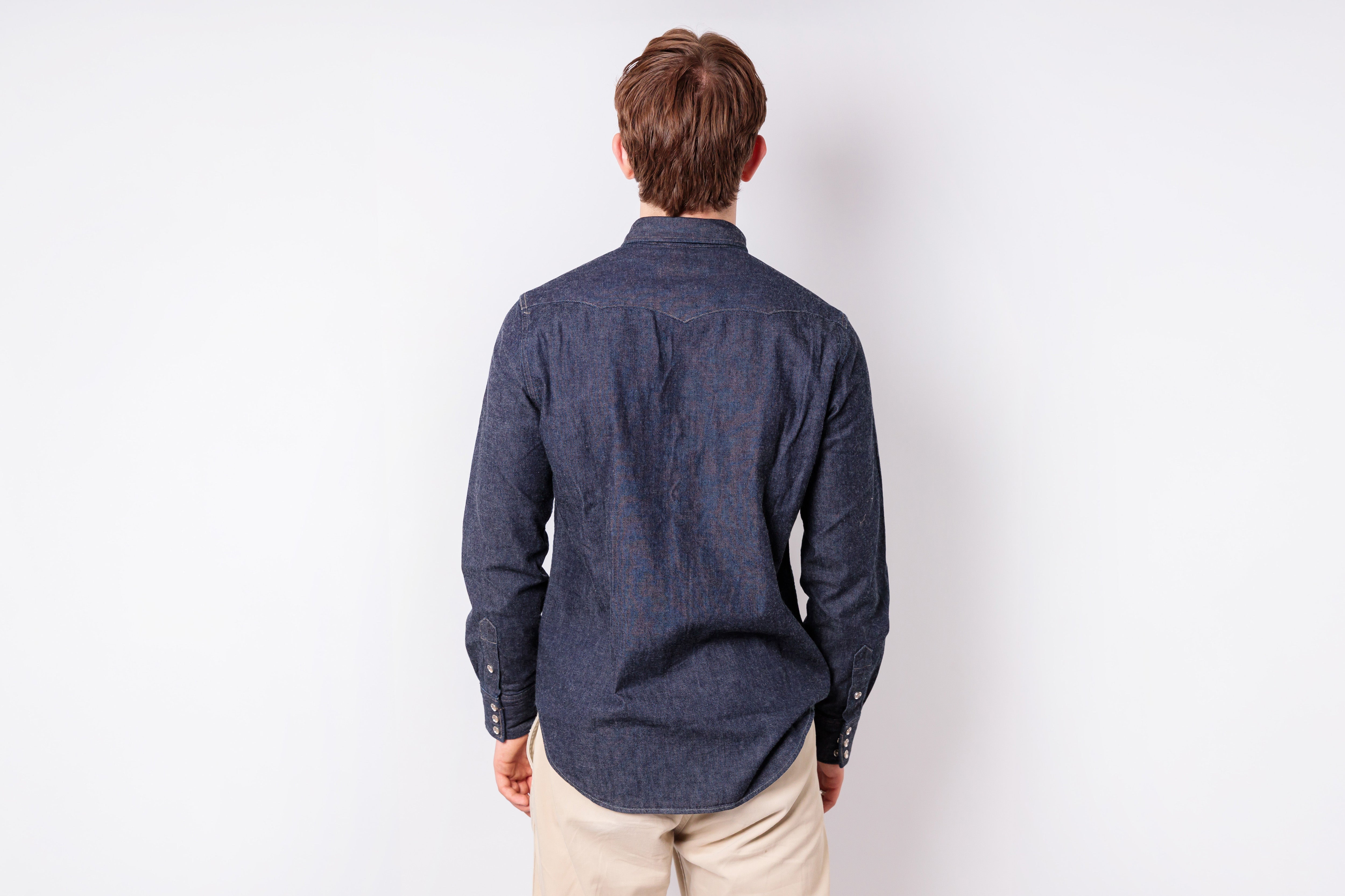 A man viewed from behind wearing a dark navy blue patterned long-sleeve button-up shirt and beige trousers, standing against a plain white studio background.