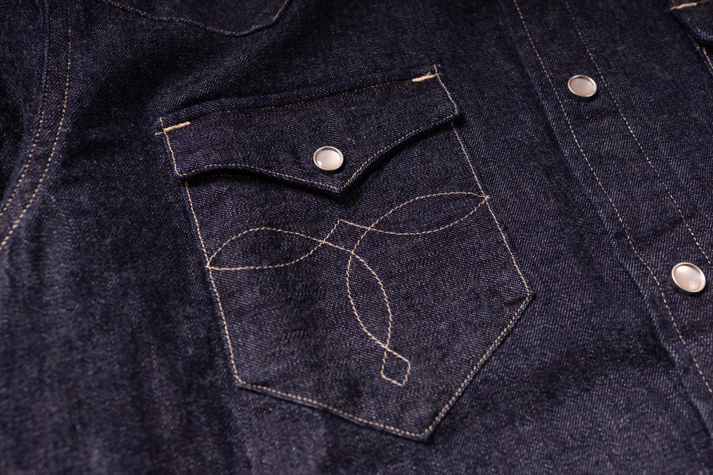 Close-up of a dark indigo denim jacket featuring a chest pocket with light tan contrast stitching and a silver snap button.