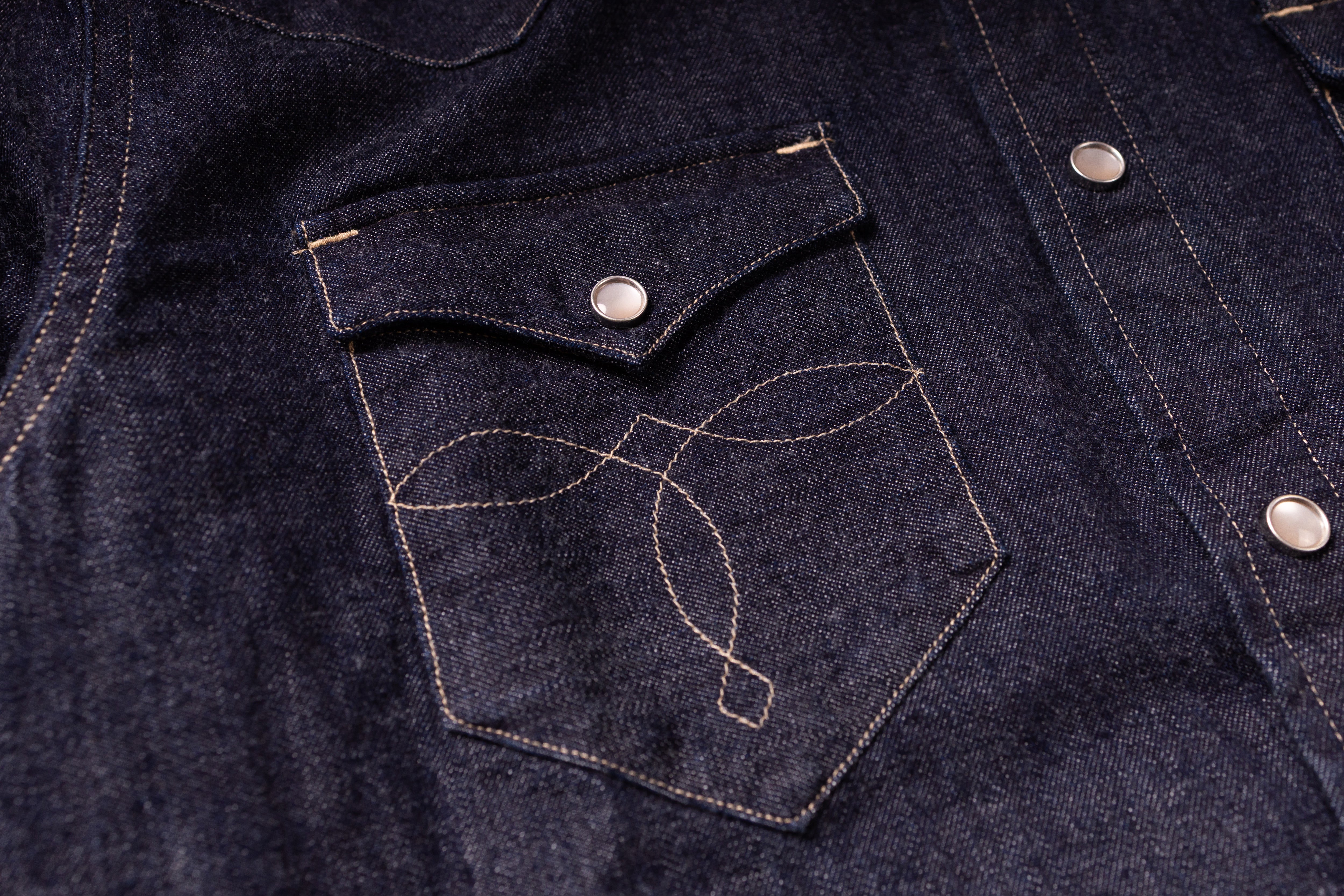 Close-up of a dark indigo denim jacket featuring a chest pocket with light tan contrast stitching and a silver snap button.