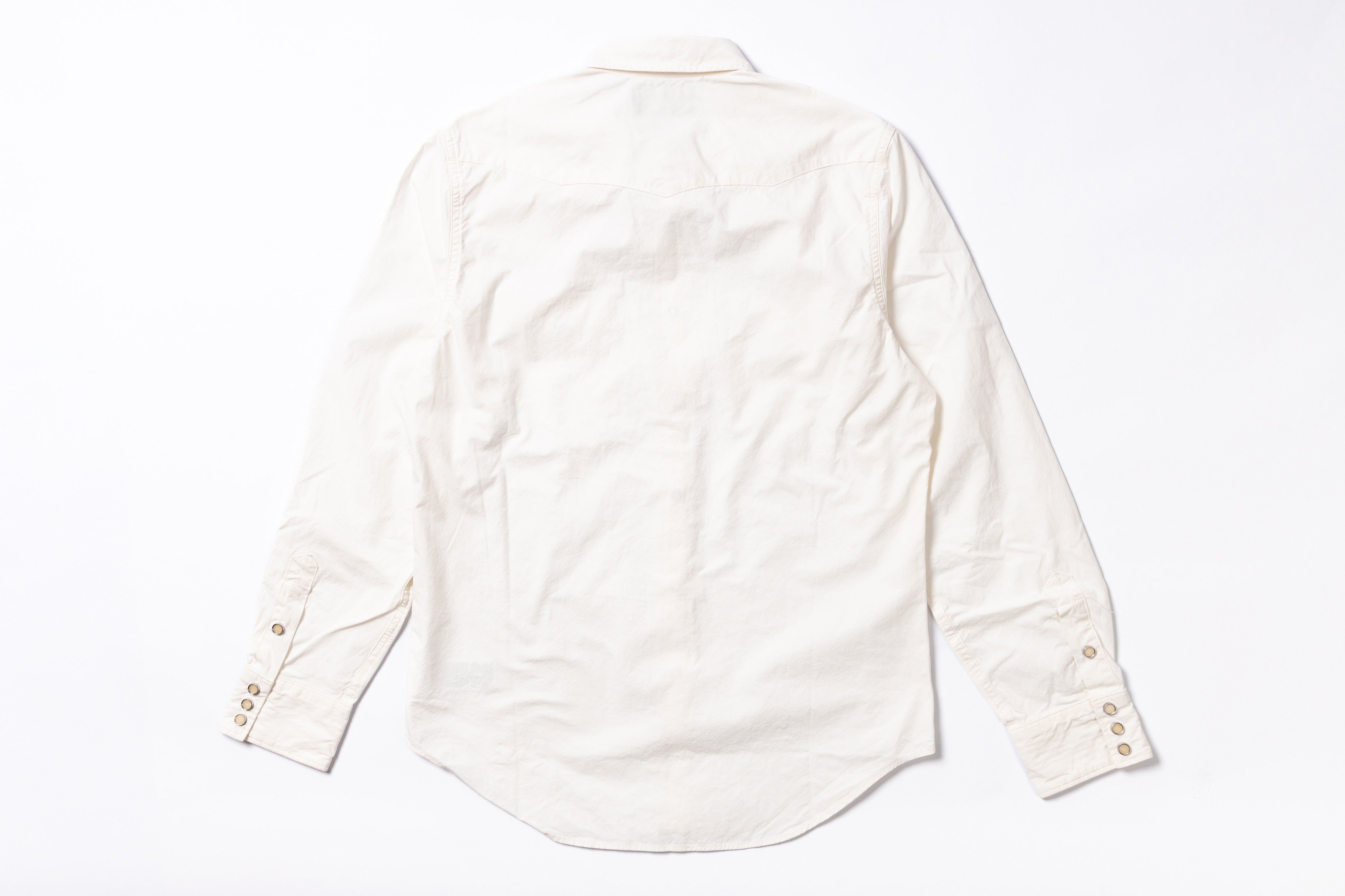 An off-white long-sleeve cotton  shirt shown from the back, laid flat on a white background. The shirt features a natural linen texture, a curved hem, and button details on the cuffs.
