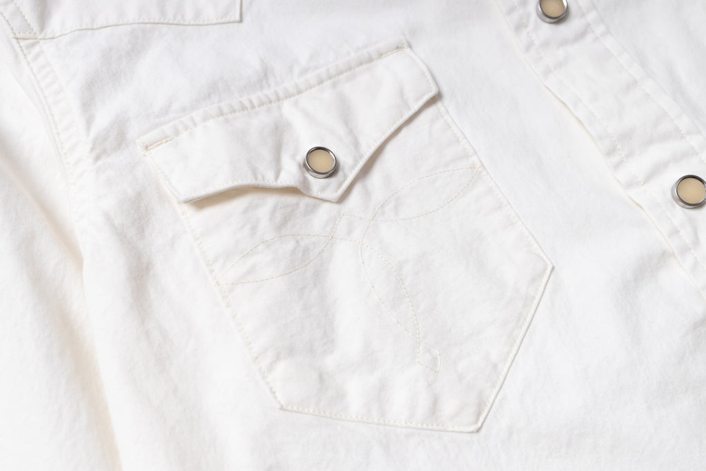 Close-up of a white button-up shirt featuring a chest pocket with a flap and decorative stitching, secured by a silver snap button.