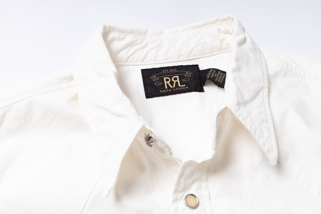 Close-up of a white button-up shirt collar with a black neck label featuring gold lettering.