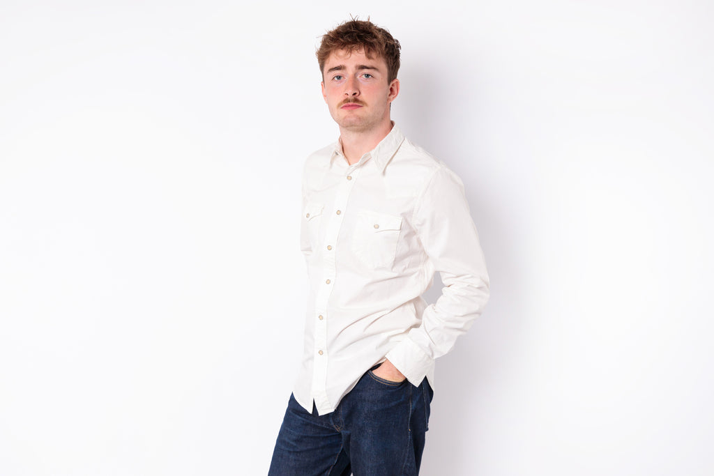 A man wearing a white long-sleeve button-up shirt and dark blue jeans, standing against a plain white studio background.