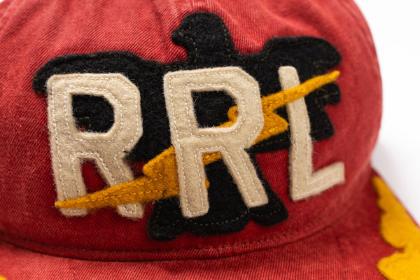 Close-up of a red baseball cap featuring a large embroidered patch with the letters 'RRL' in cream, outlined in black, with a yellow lightning bolt design.