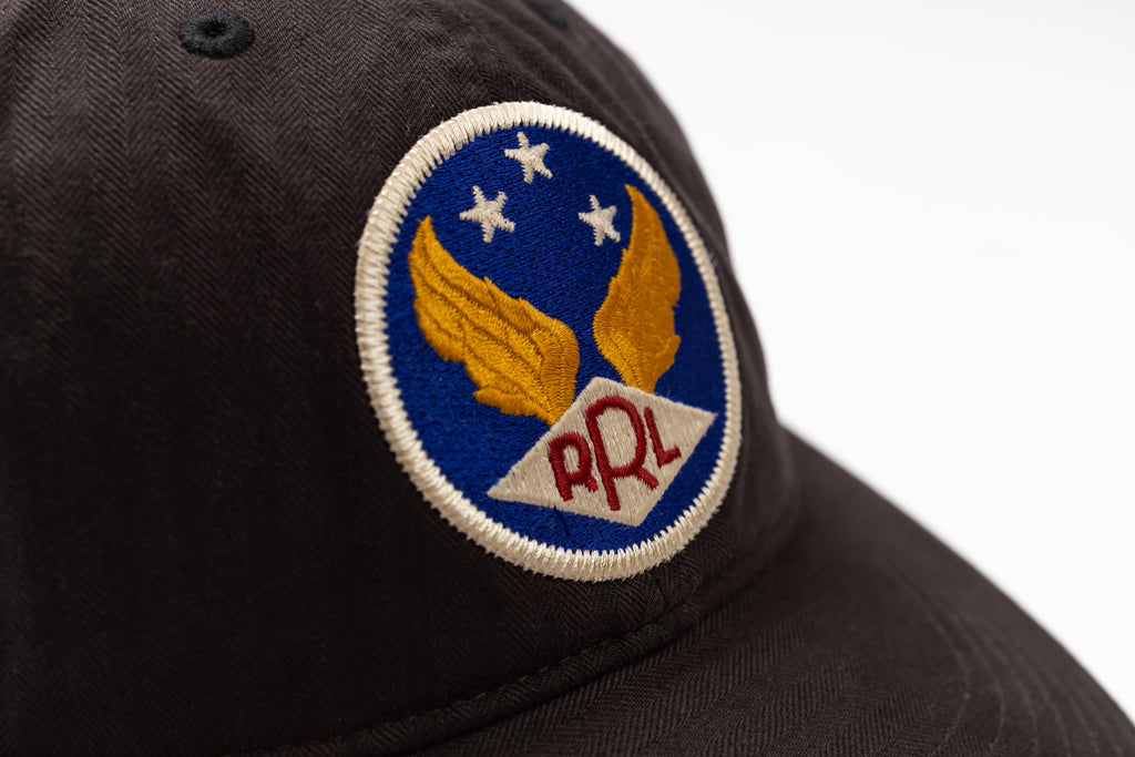 Close-up of a dark brown or black baseball cap featuring a circular embroidered patch with gold wings, white stars on a blue background, and red 'RRL' lettering on a white banner.