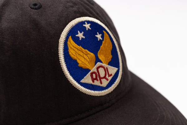 Close-up of a dark brown or black baseball cap featuring a circular embroidered patch with gold wings, white stars on a blue background, and red 'RRL' lettering on a white banner.