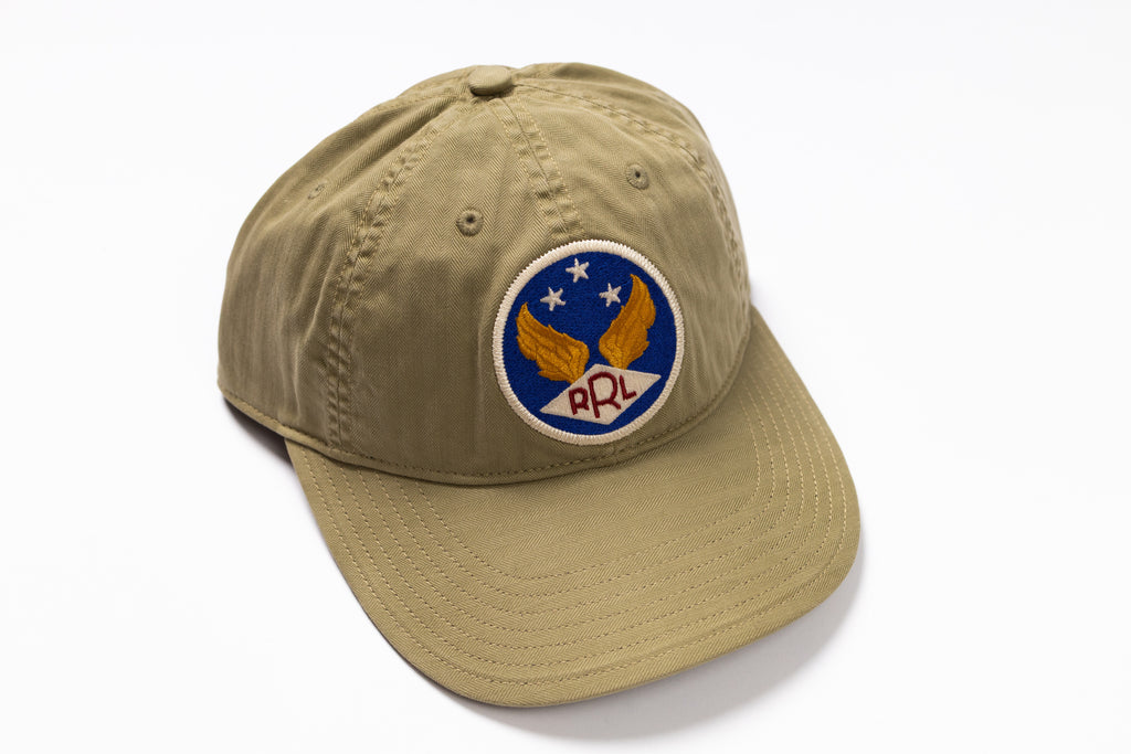 A khaki baseball cap featuring a circular embroidered patch on the front with wings, stars, and the letters 'RRL' on a white background.