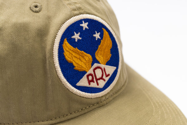 Close-up of a khaki baseball cap featuring a circular embroidered patch with a blue background, white stars, yellow wings, and the letters 'RRL' in red.