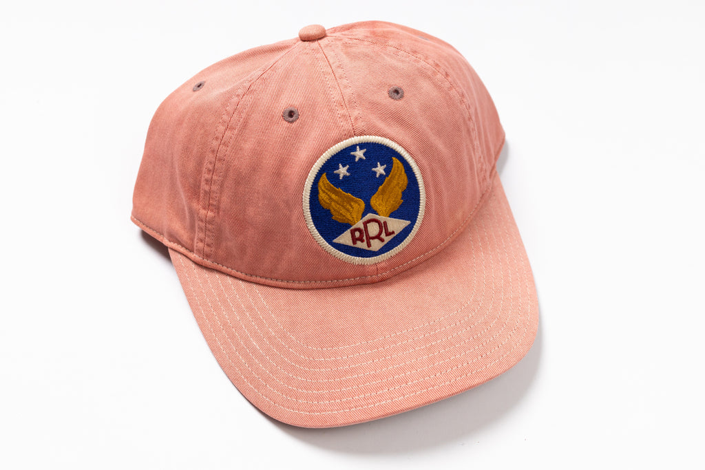 A washed peach-pink baseball cap featuring a circular embroidered patch on the front with a blue background, gold wings, white stars, and the letters 'RRL' in red.