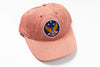 A washed peach-pink baseball cap featuring a circular embroidered patch on the front with a blue background, gold wings, white stars, and the letters 'RRL' in red.