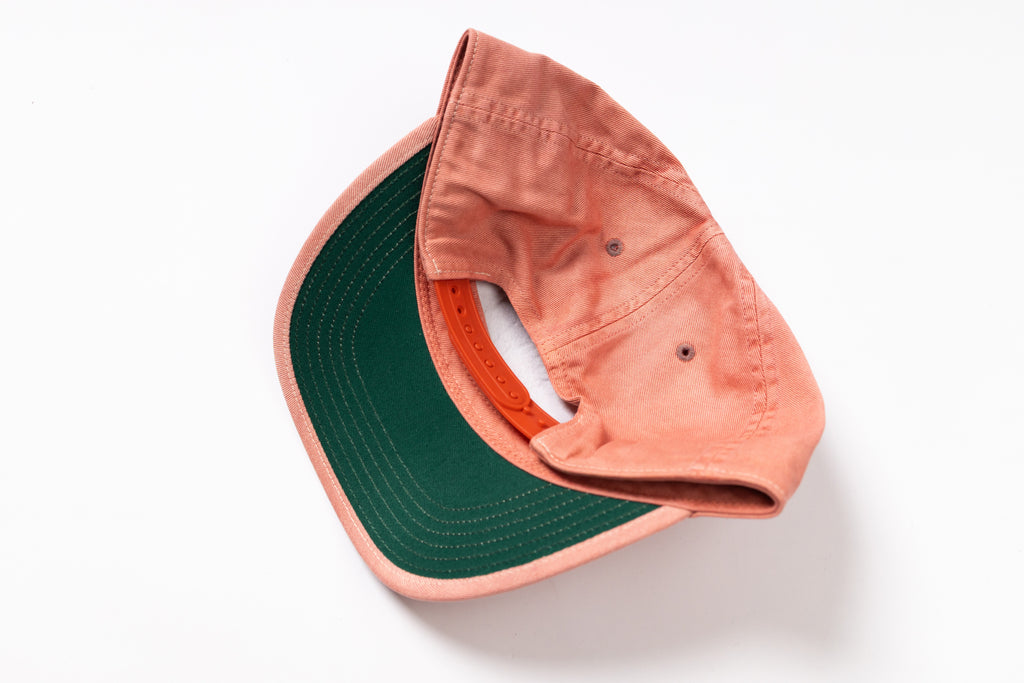 A folded baseball cap shown from the back, featuring a dusty coral-pink fabric crown and a dark forest green visor, photographed on a clean white background.