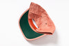 A folded baseball cap shown from the back, featuring a dusty coral-pink fabric crown and a dark forest green visor, photographed on a clean white background.