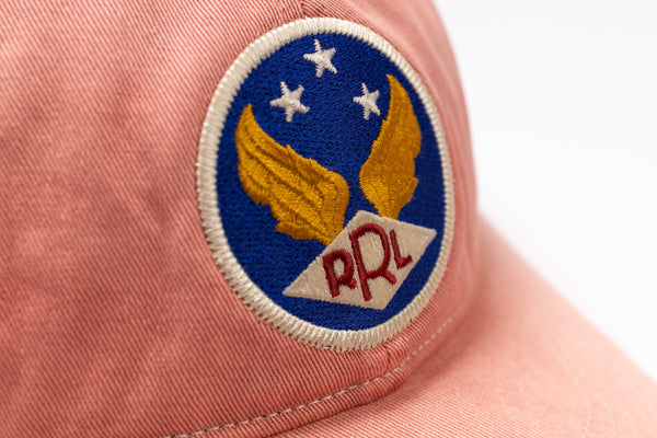 Close-up of a dusty pink cap featuring a circular embroidered patch with a blue background, gold wings, three white stars, and red 'RRL' lettering on a white diamond.