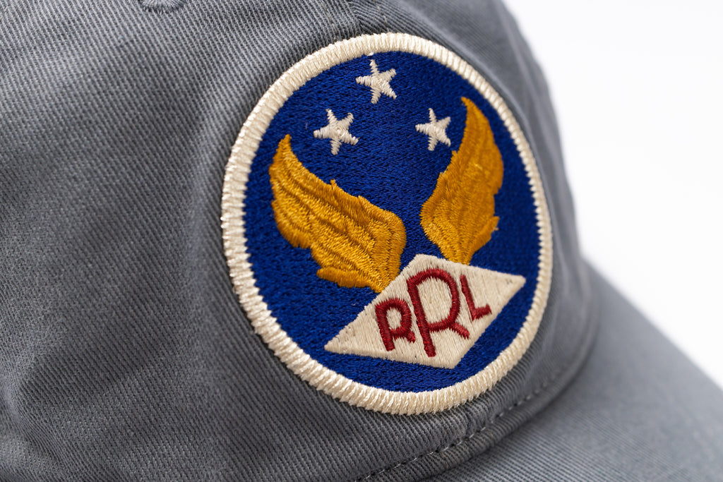 Close-up of a gray baseball cap featuring a circular embroidered patch with gold wings, three white stars on a blue background, and red 'RRL' lettering on a white diamond.