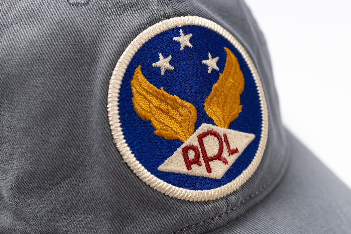 Close-up of a gray baseball cap featuring a circular embroidered patch with gold wings, three white stars on a blue background, and red 'RRL' lettering on a white diamond.