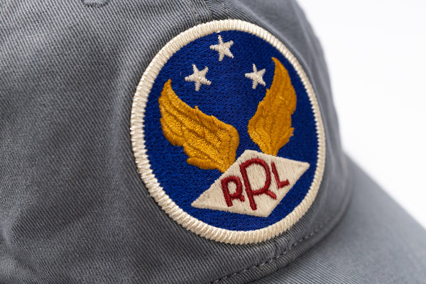 Close-up of a gray baseball cap featuring a circular embroidered patch with gold wings, three white stars on a blue background, and red 'RRL' lettering on a white diamond.