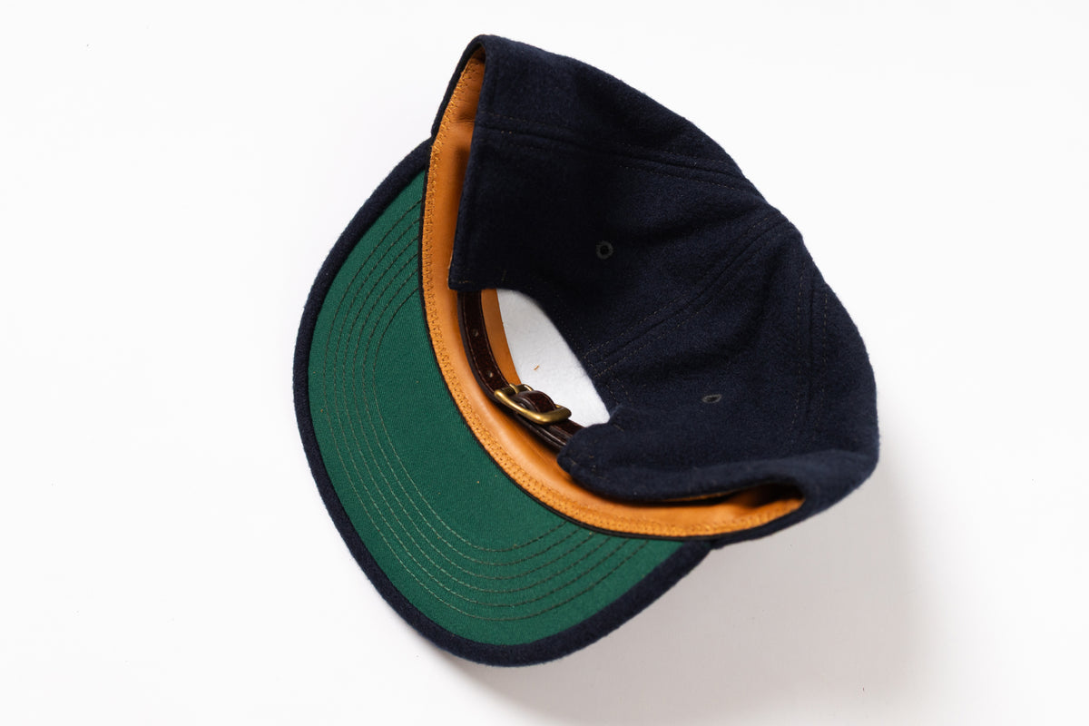 A dark navy blue baseball cap with a green visor and a tan leather-like trim along the edge, shown from an overhead angle on a white background.