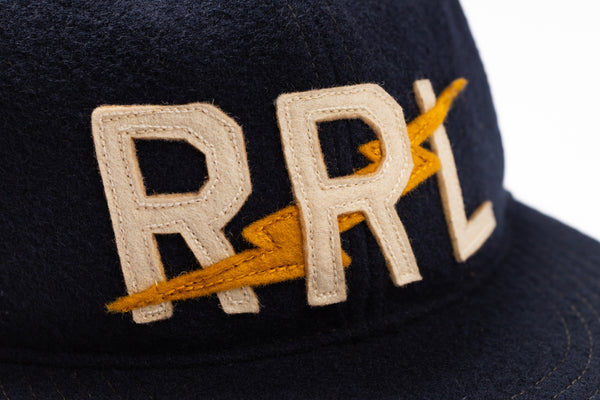 Close-up of a dark navy baseball cap featuring cream-colored embroidered letters 'RRL' with a yellow lightning bolt graphic.