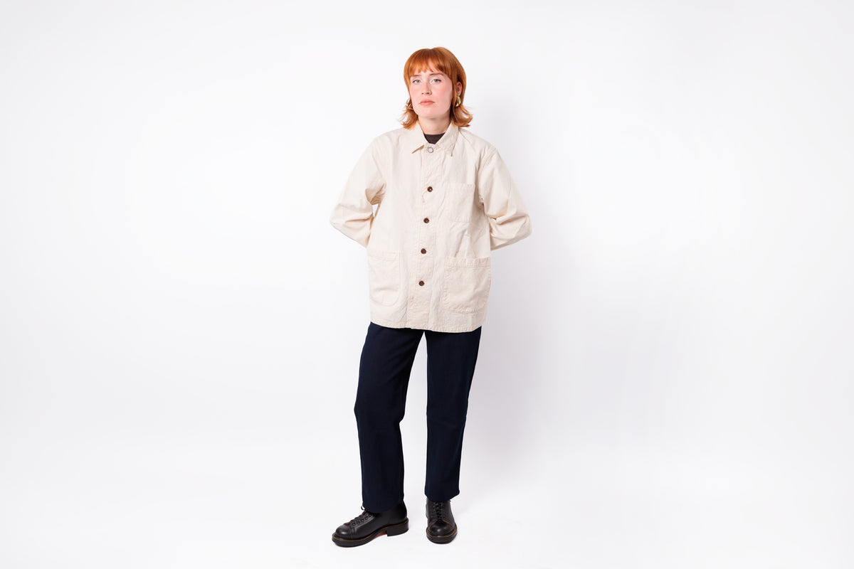 A person with red hair wearing a cream-colored button-up shirt jacket, dark navy pants, and black shoes, standing against a plain white studio background.