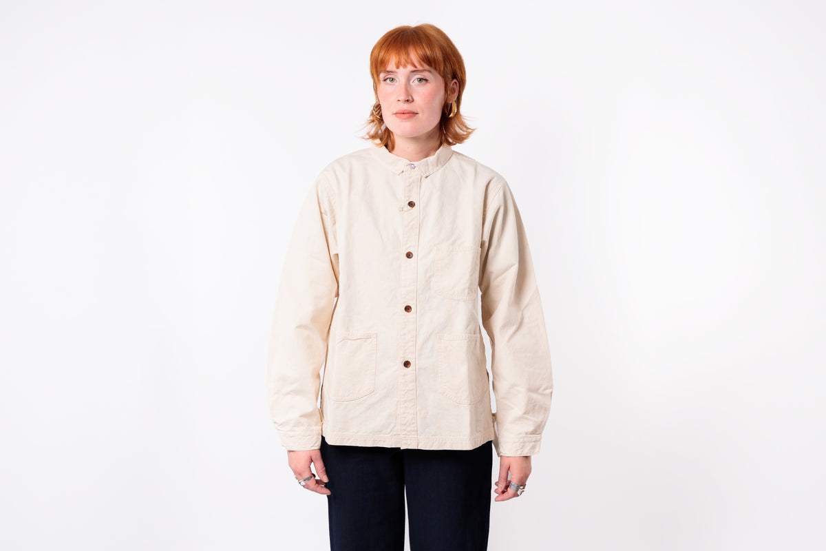 A model with short red hair wearing a light beige, long-sleeved button-up shirt jacket and dark pants, standing against a plain white studio background.