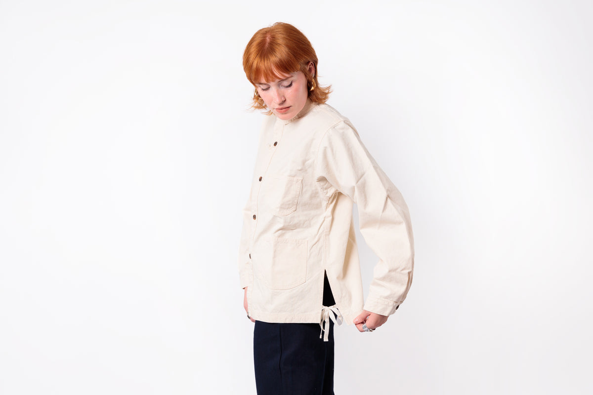A model with red hair wearing a cream-colored, button-up linen jacket and dark trousers, standing against a plain white studio background.