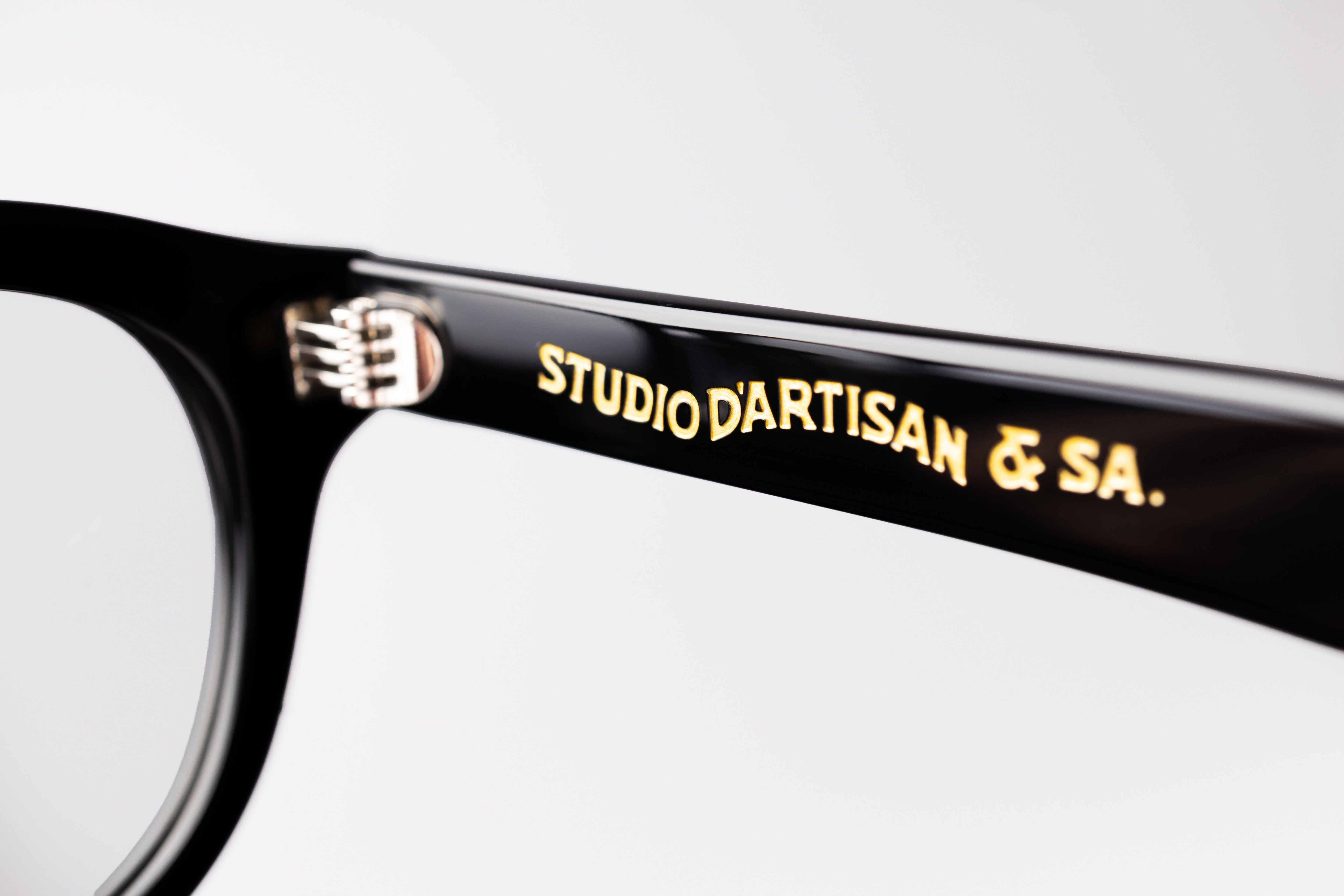 Close-up of a black eyeglass frame with 'Studio d'Artisan & SA.' engraving on a white background