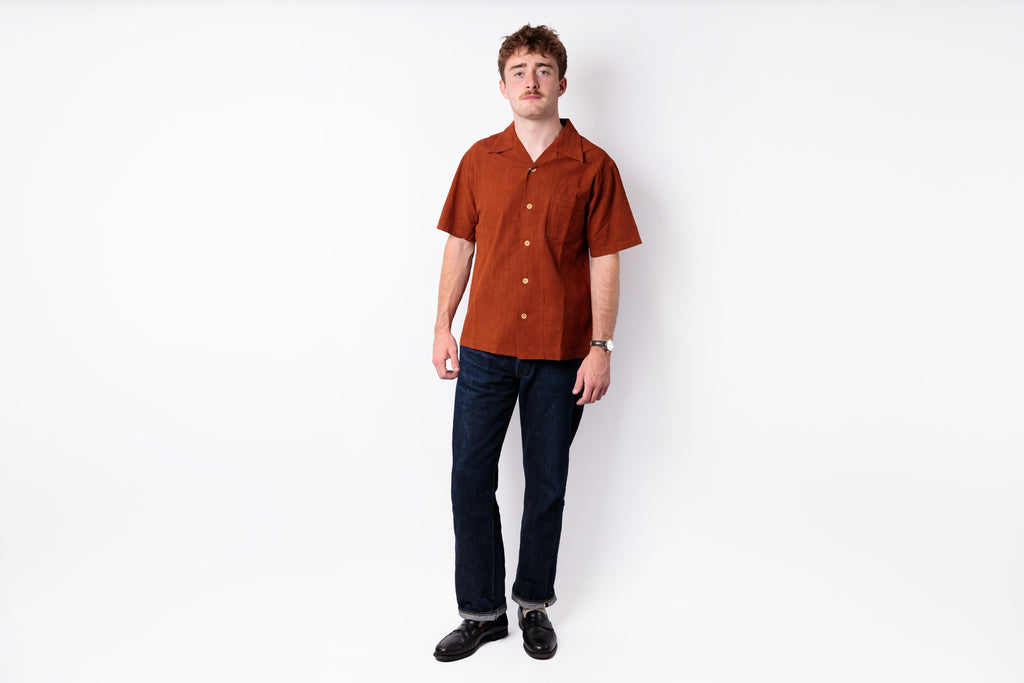 A man modeling a short-sleeve button-up shirt in rust orange, paired with dark blue jeans and black shoes, standing against a plain white studio background.